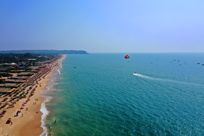Aerial view of Candolim Beach North Goa with beach to the left and ocean to the right. Above is someone with a multicoloured parasail being towed by a motor boat.